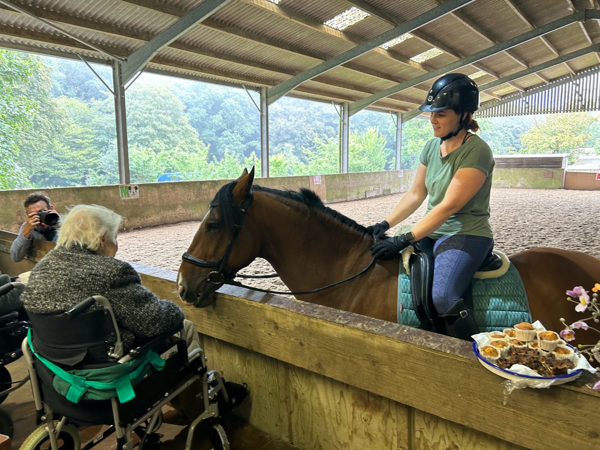An elderly person in a wheelchair in the indoor arena reaches out to a horse ridden by a volunteer. Cakes are laid out on the arena fence nearby.