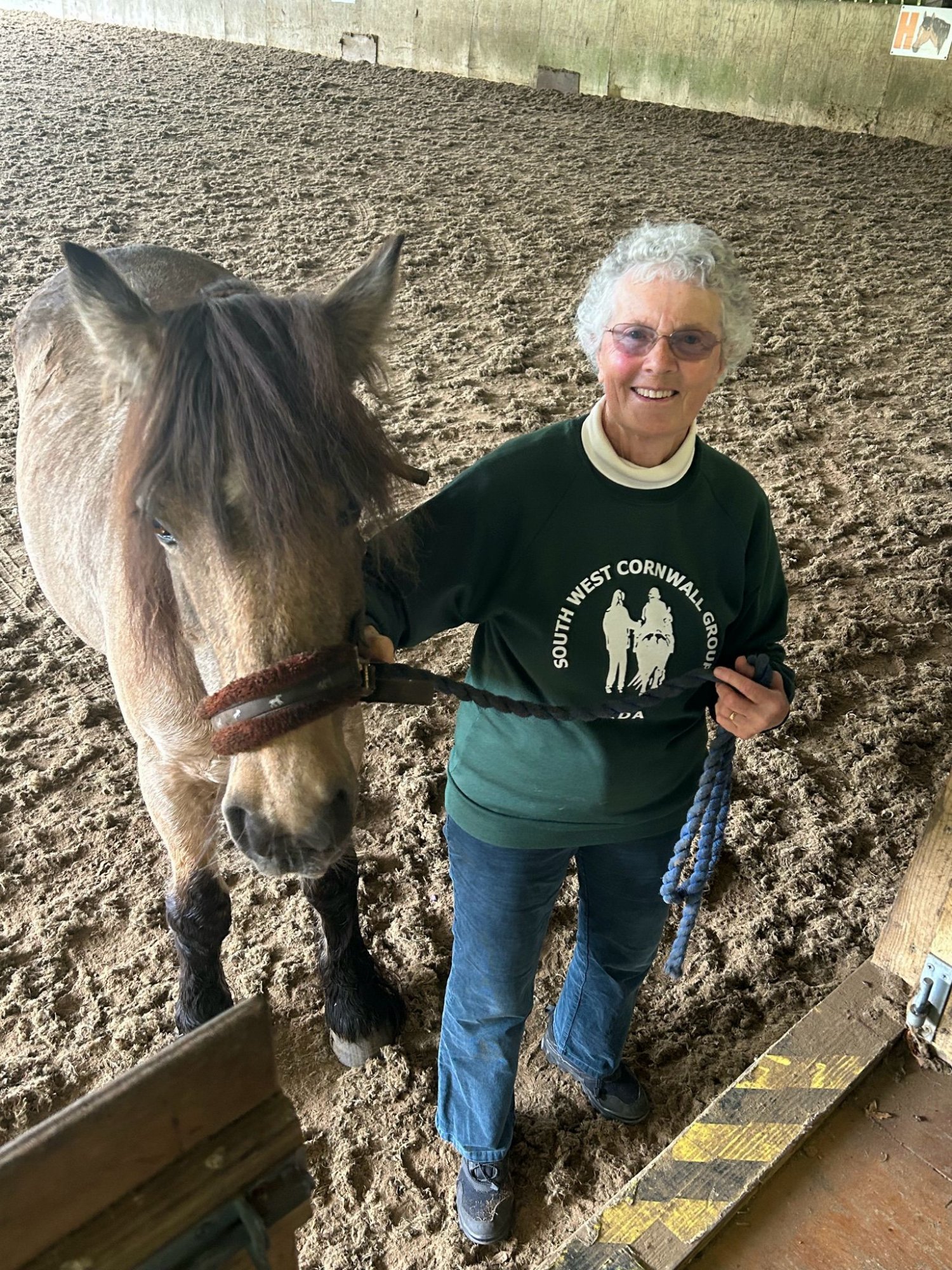 A smiling volunteer wearing a South West Cornwall Group RDA sweatshirt stands beside a grey horse in the indoor arena
