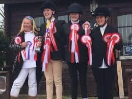 Four young riders smiling proudly in front of the stables, each holding large red and white competition rosettes