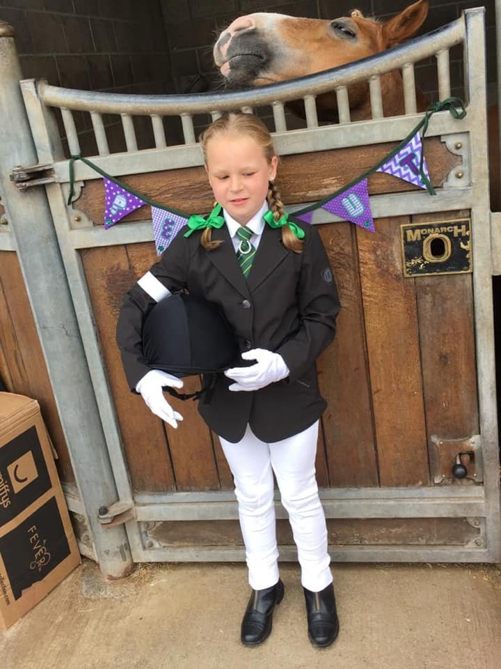 Young rider in smart competition clothing holding a riding hat, standing in front of a stable door with a horse looking over