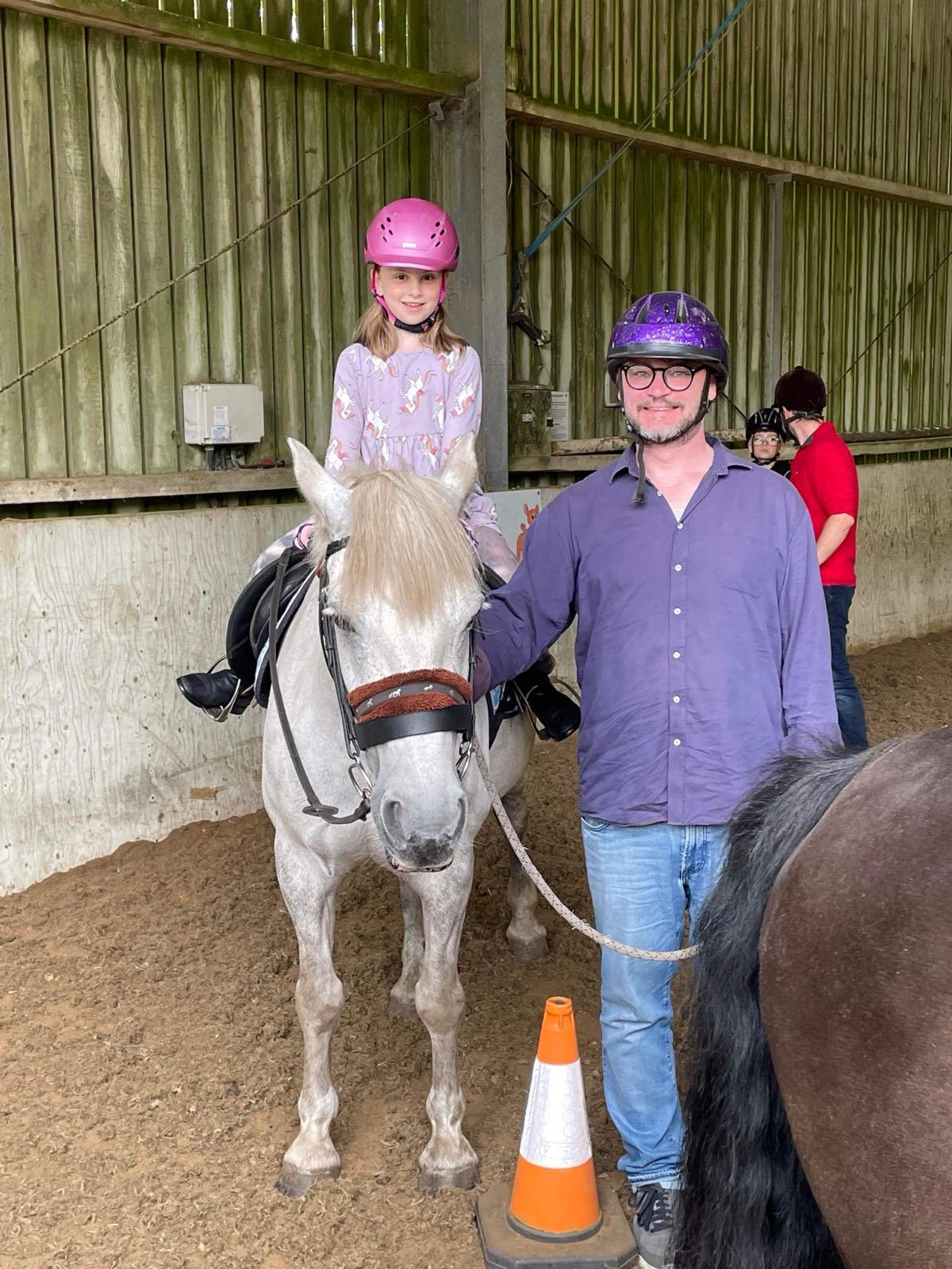 A young rider in a pink helmet sits on Rose, our grey RDA horse, with a supporter standing alongside in the indoor arena